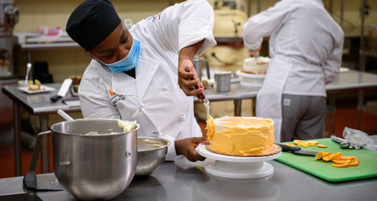 female baking student preparing a cake in a commercial kitchen with another student baker in the background